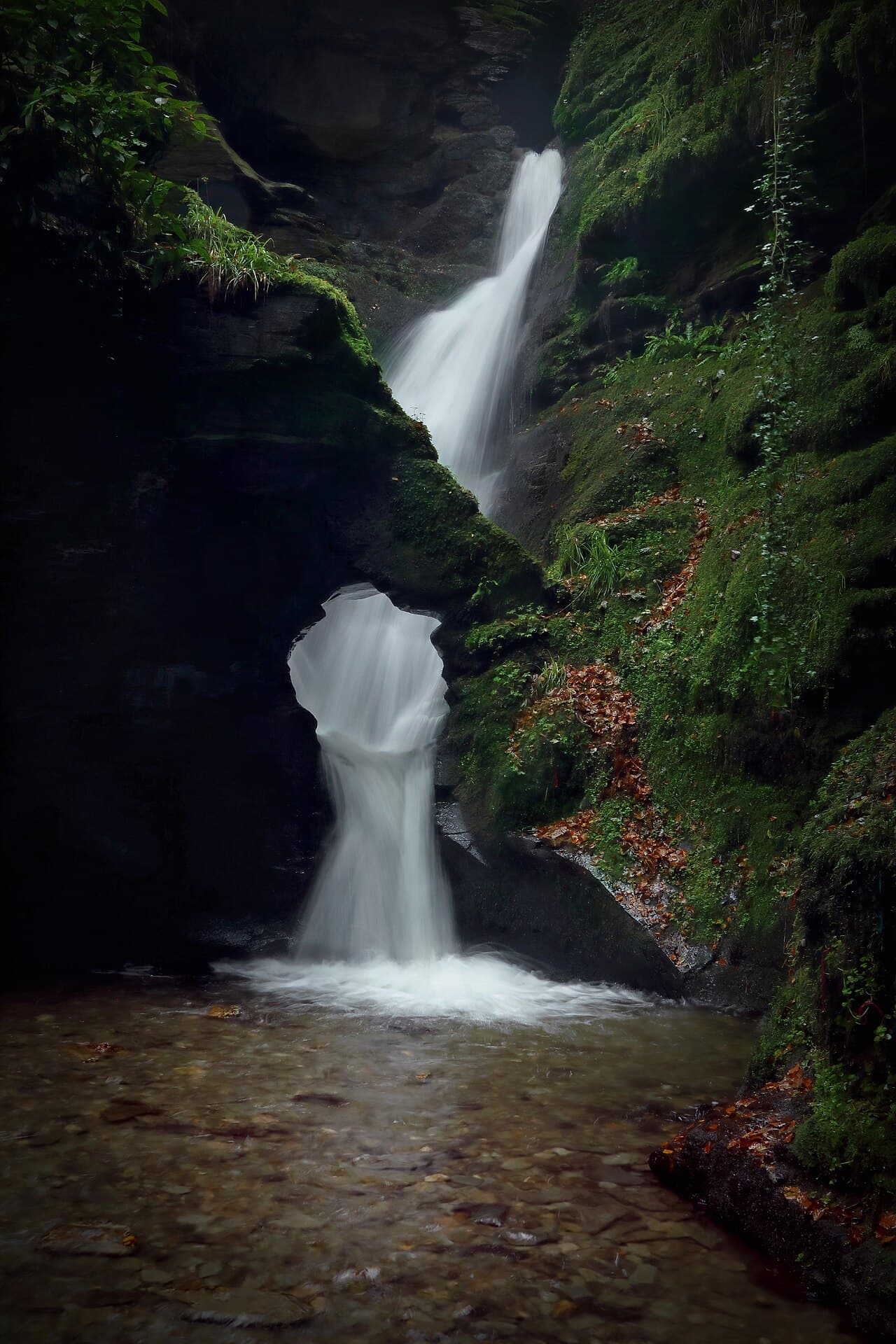 The waterfall at St Nectan's Kieve pouring through a circular hole in dark slate, long-exposure water falling silky-white past moss and scattered autumn leaves into a shallow pebbled pool.