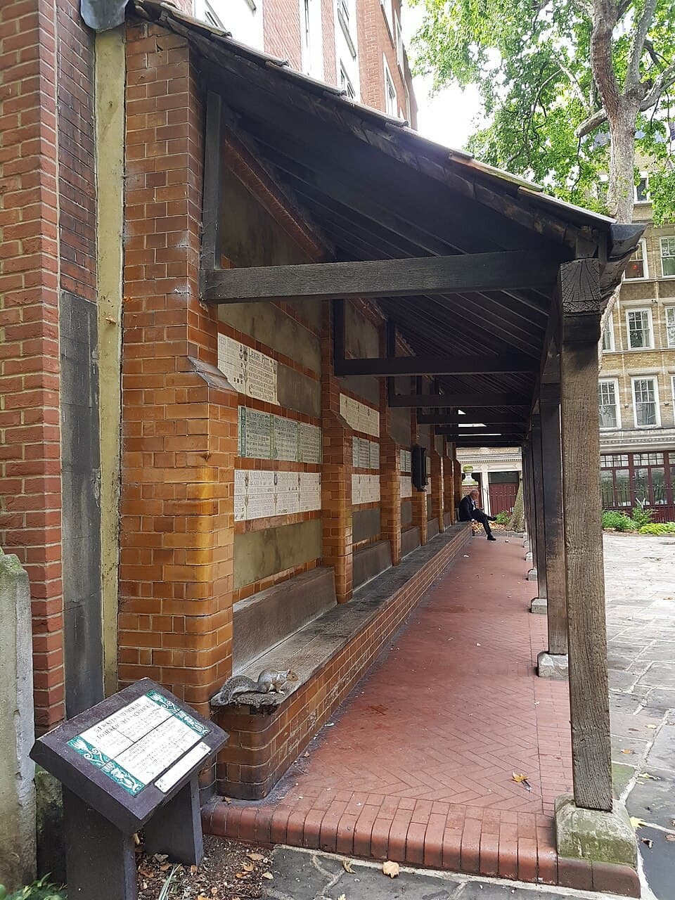 View down the timber-roofed loggia of the Watts Memorial, its brick wall lined with glazed ceramic tablets, a squirrel perched on the long bench and a lone visitor seated at the far end.