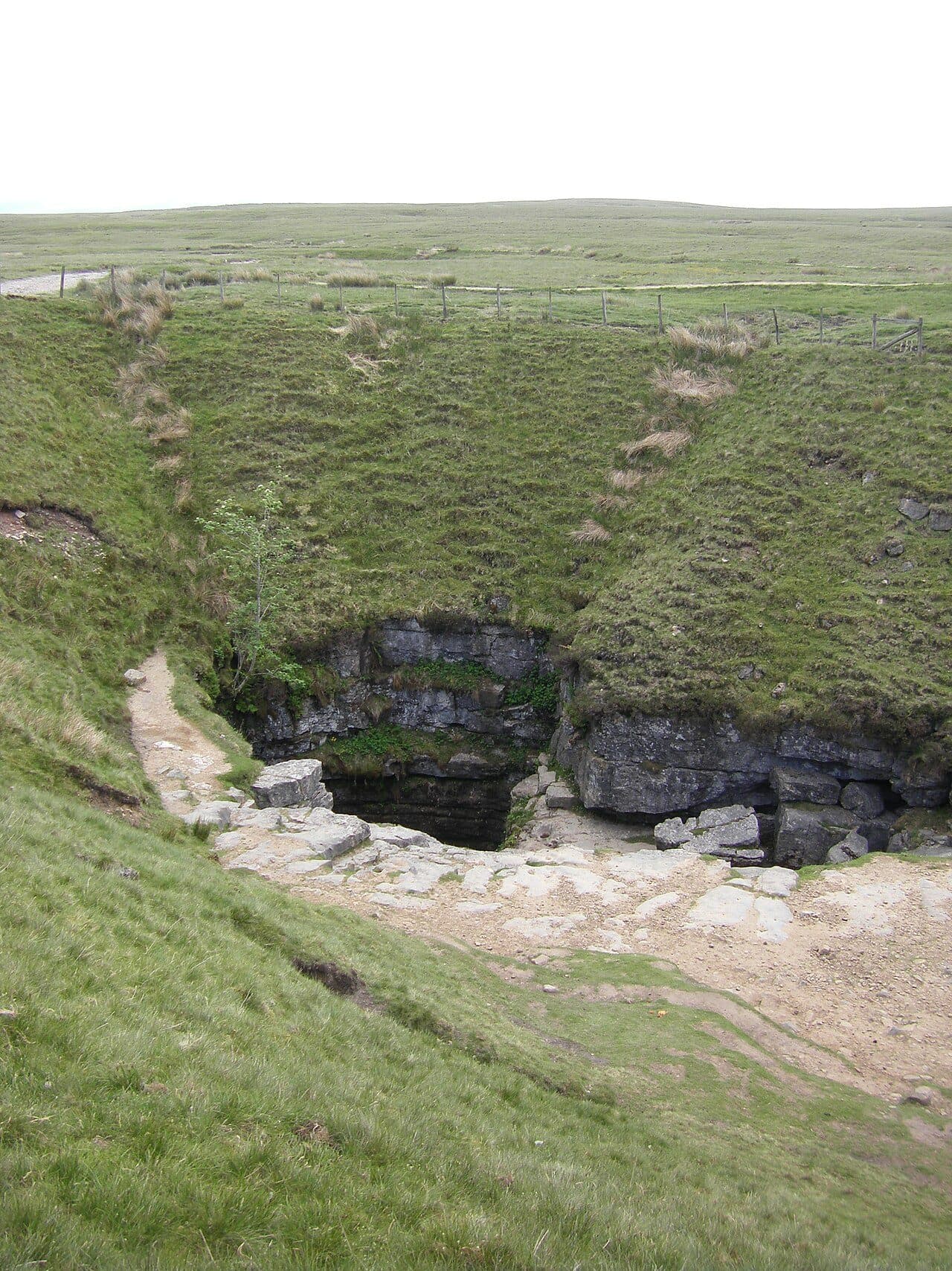 The dark mouth of Gaping Gill opens beneath a limestone lip in a grassy hollow of Ingleborough's moor, a worn path winding down to its edge.